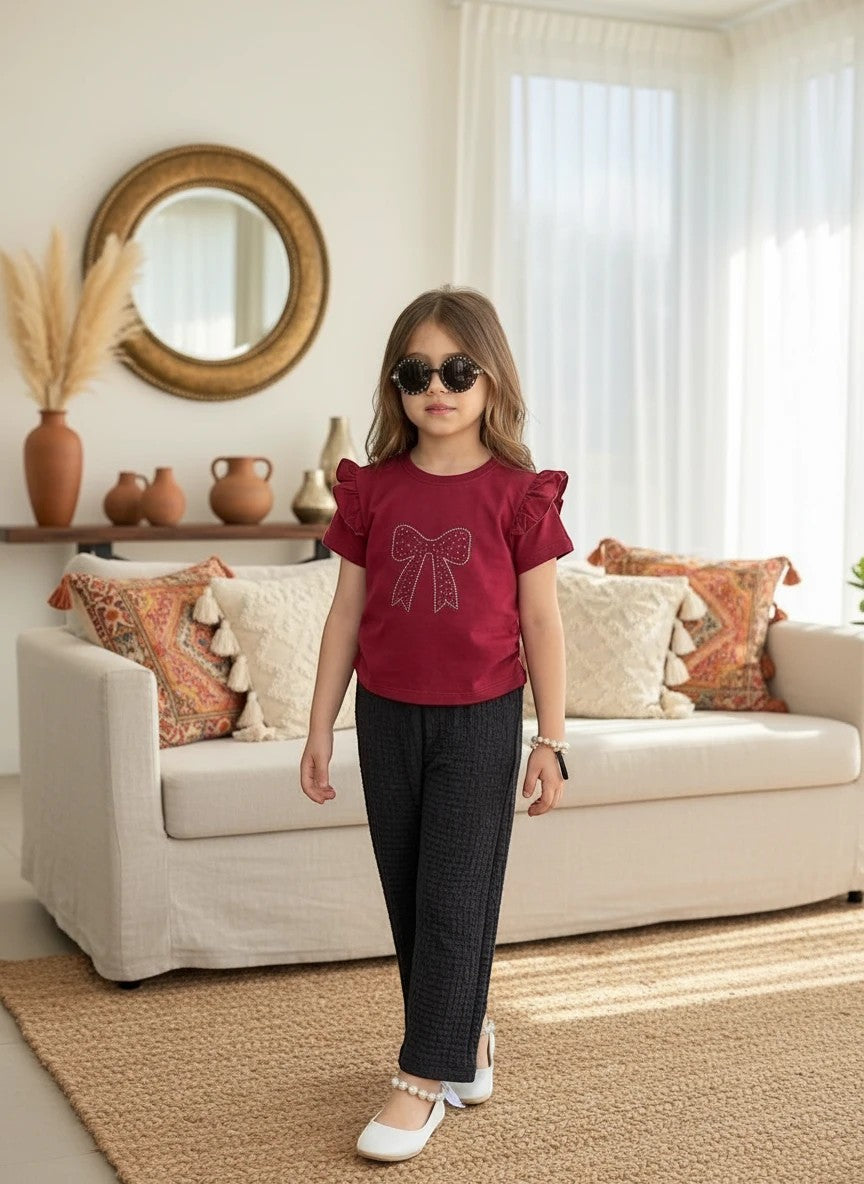 Girl wearing The Nile Luxuries burgundy ruffle sleeve top with sparkling bow and black textured trousers in a living room