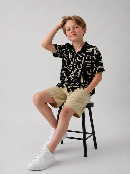 The Nile Luxuries black short sleeve shirt with abstract print, collared neckline, worn by a boy sitting on a stool.