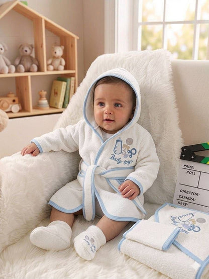 The Nile Luxuries baby wearing a white hooded bathrobe with blue trim and baby toy embroidery, sitting on a chair.