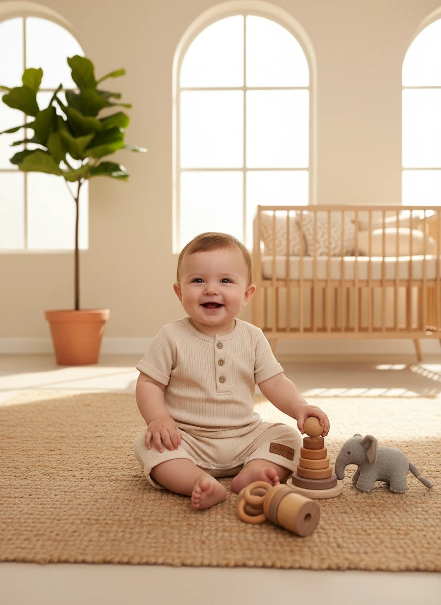 A happy baby wearing a beige ribbed cotton romper by The Nile Luxuries, sitting on a rug with wooden toys.