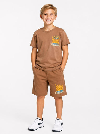 A smiling boy wearing a brown The Nile Luxuries cotton t-shirt and shorts set with a retro California beach graphic.