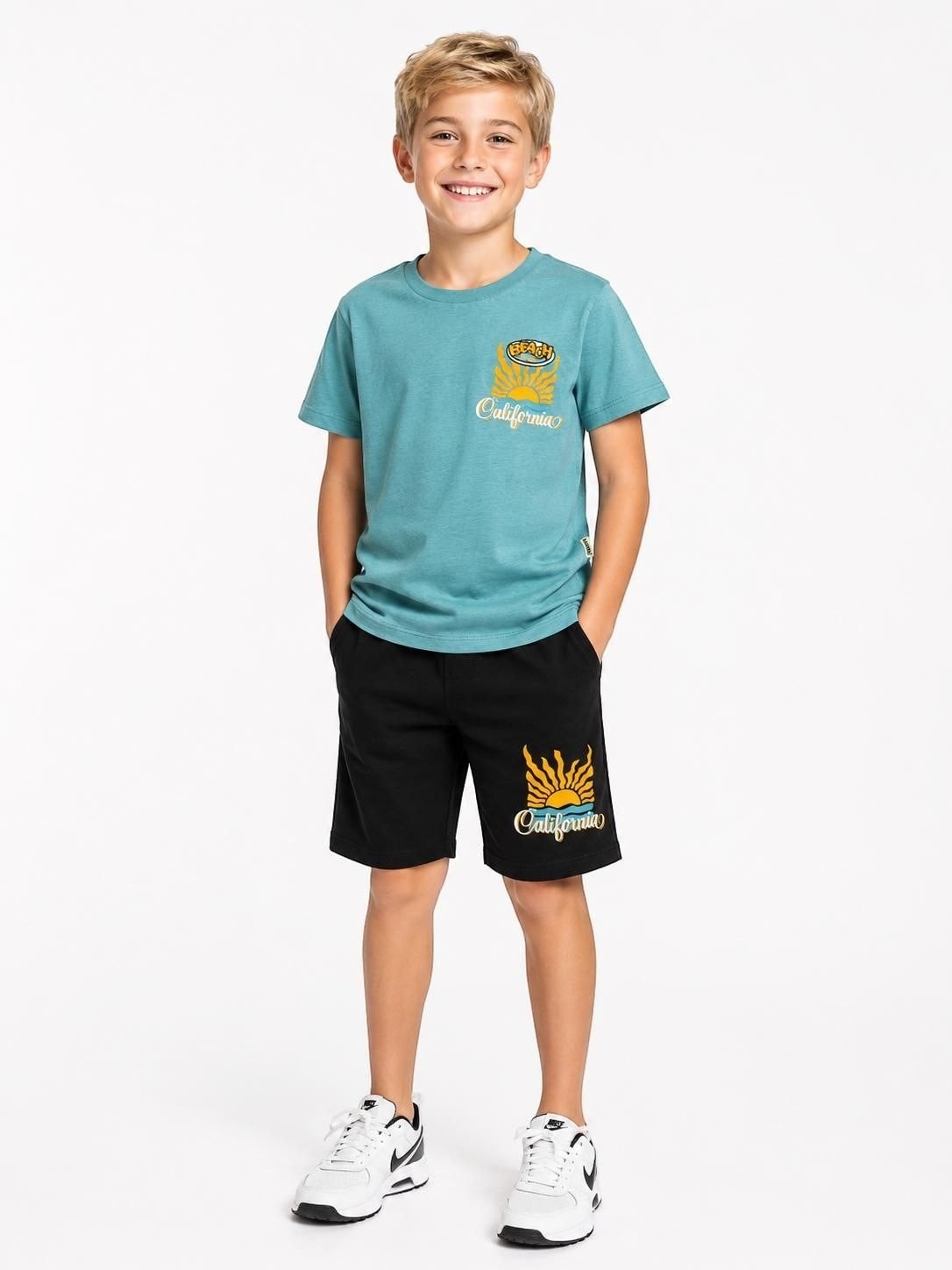 A young boy wearing a The Nile Luxuries teal crew-neck t-shirt and black shorts with California beach graphic prints.
