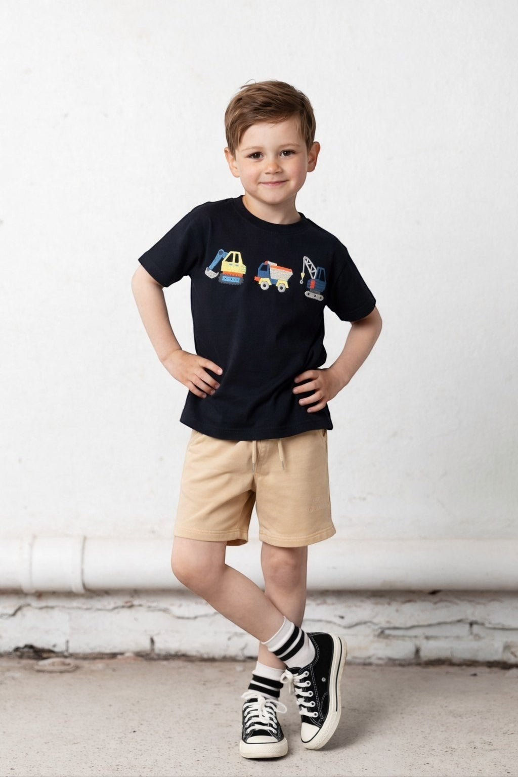 A young boy wearing The Nile Luxuries navy t-shirt with construction vehicle embroidery and beige pull-on shorts.