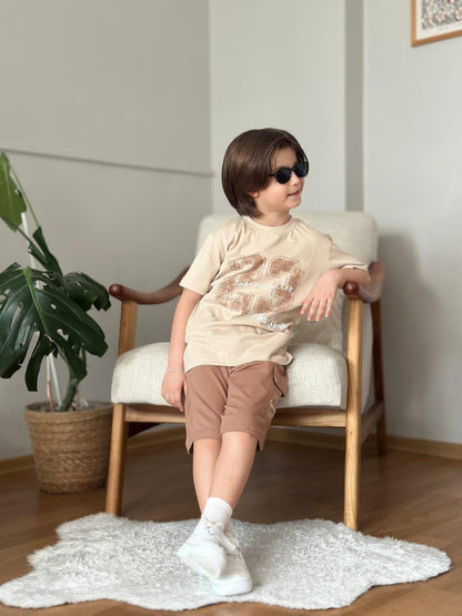 A boy wearing The Nile Luxuries beige graphic t-shirt and brown cargo shorts, sitting on a chair with sunglasses.