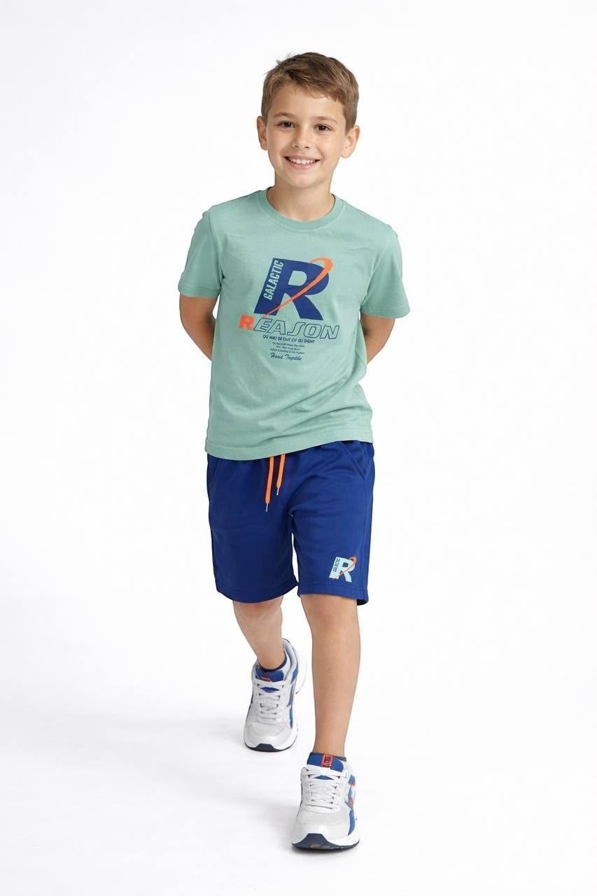 A young boy wearing a The Nile Luxuries sage green graphic t-shirt and blue athletic shorts with an orange drawstring.