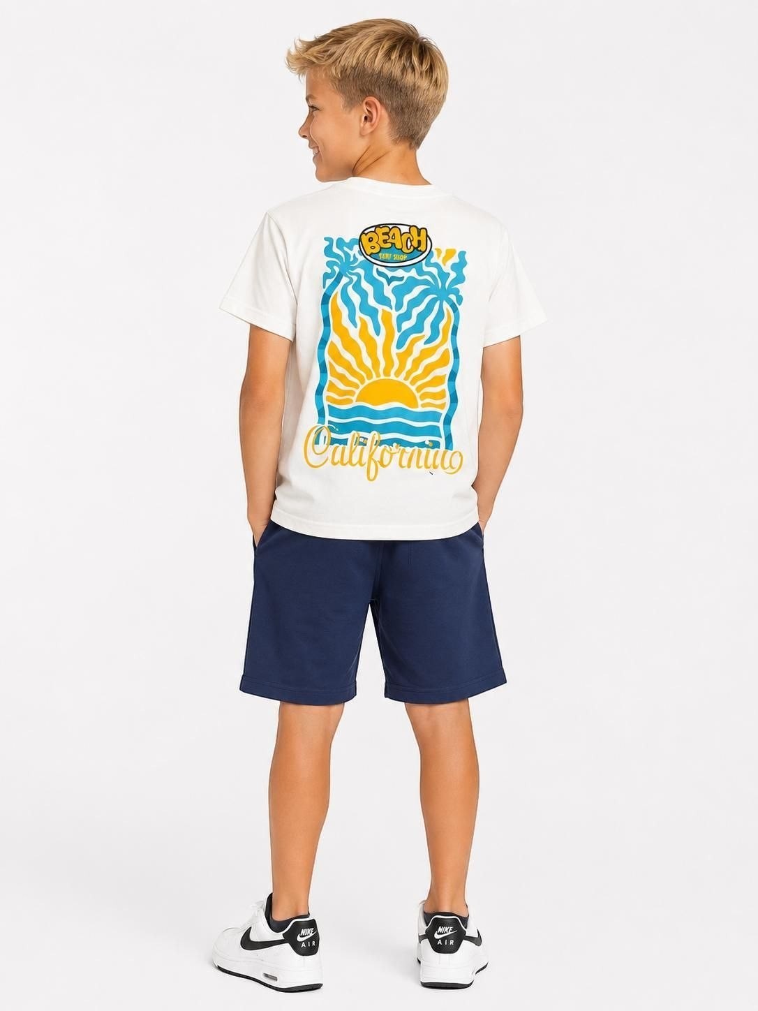 Boy wearing The Nile Luxuries white t-shirt with California beach graphic and navy blue shorts against white background