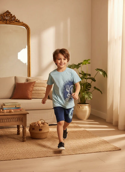 A young boy wearing a The Nile Luxuries light blue graphic t-shirt and navy cargo shorts in a bright living room.