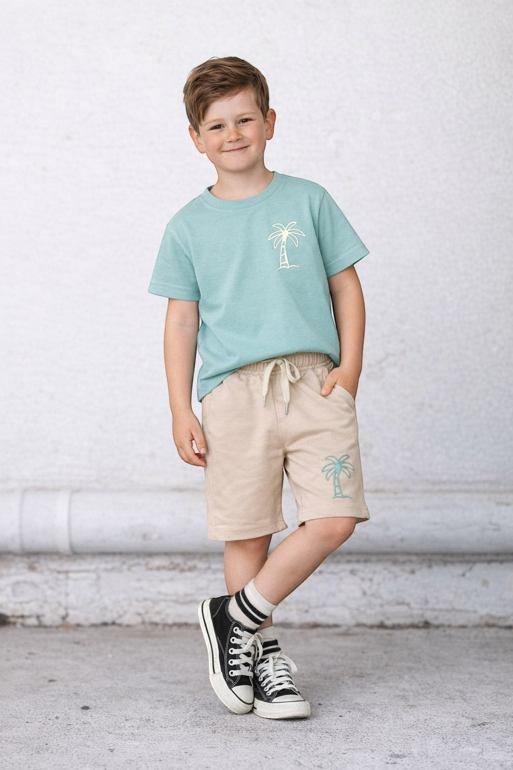 The Nile Luxuries boy wearing a sage green palm tree t-shirt and beige drawstring shorts for a casual summer look.