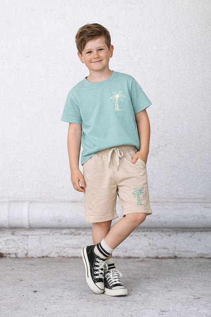 The Nile Luxuries boy wearing a sage green palm tree t-shirt and beige drawstring shorts for a casual summer look.
