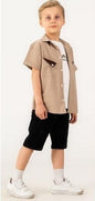 The Nile Luxuries boys' short-sleeved beige shirt with camp collar and triangular pocket detail, worn over a white t-shirt and black shorts.
