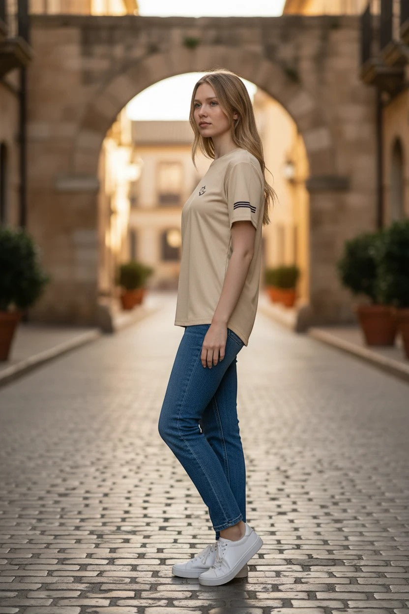 The Nile Luxuries cream performance t-shirt with sleeve stripes worn by a woman in a cobblestone street setting.