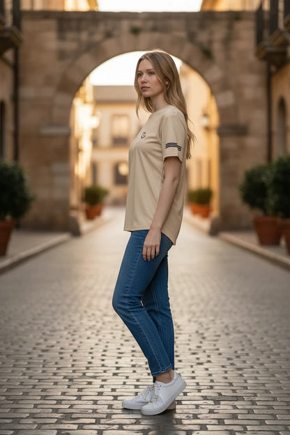 The Nile Luxuries cream performance t-shirt with sleeve stripes worn by a woman in a cobblestone street setting.