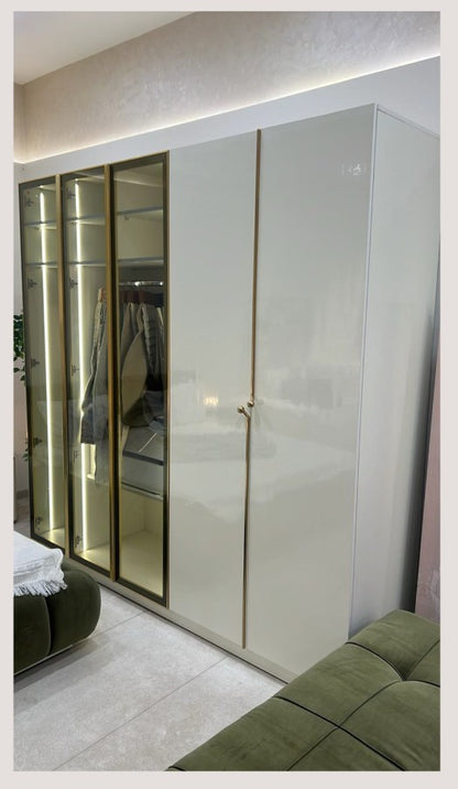 The Nile Luxuries high-gloss cream wardrobe with gold hardware and glass doors, featuring integrated lighting. Includes olive green ottoman.