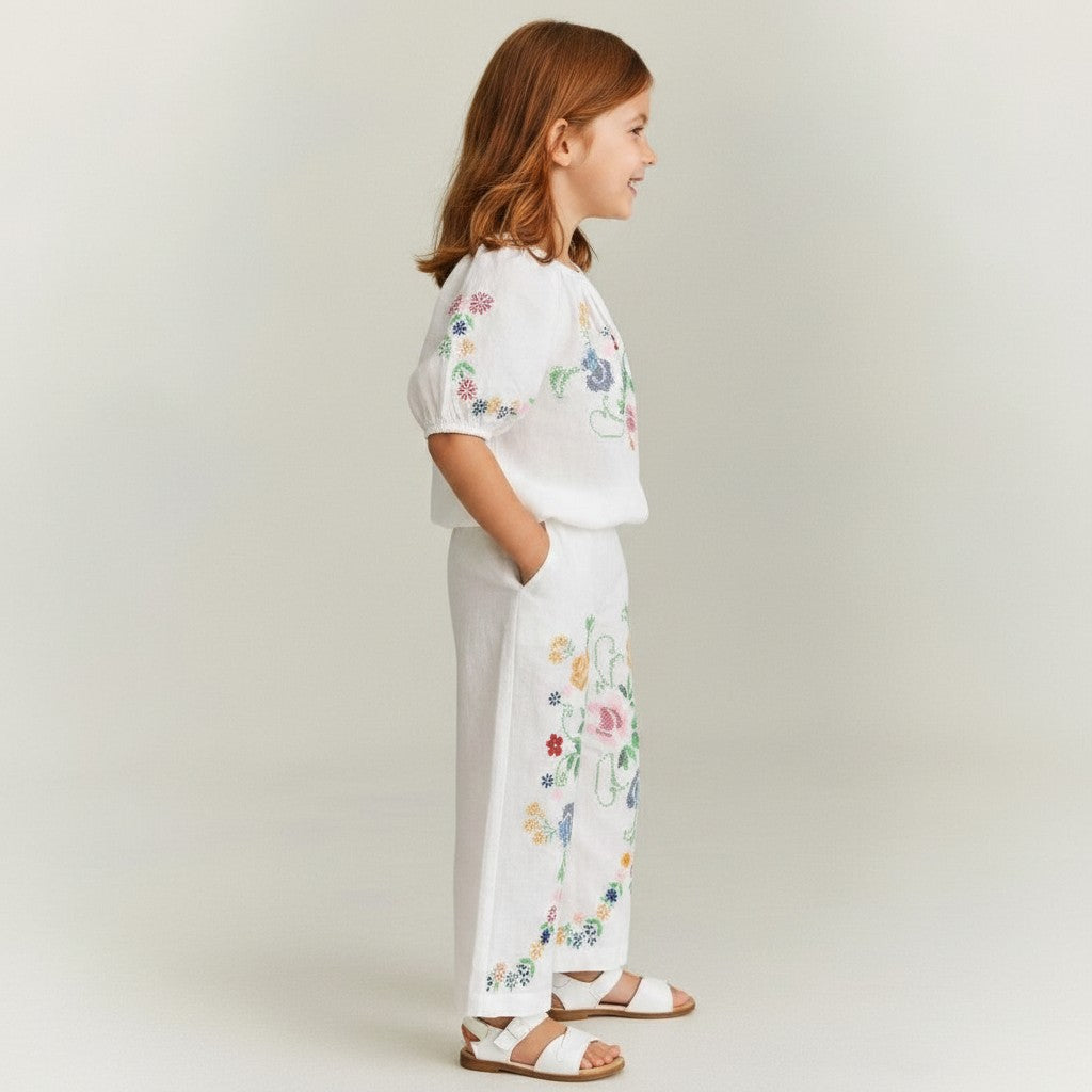 The Nile Luxuries white linen blend set for girls: off-shoulder cropped top with puff sleeves and wide-leg pants, both with colorful floral cross-stitch embroidery.