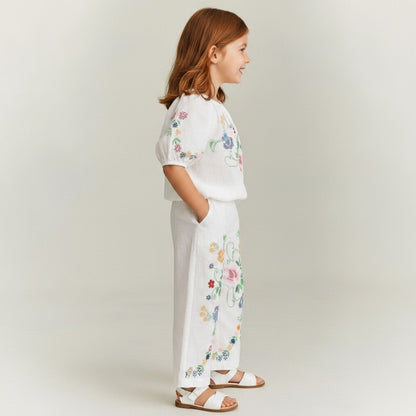 The Nile Luxuries white linen blend set for girls: off-shoulder cropped top with puff sleeves and wide-leg pants, both with colorful floral cross-stitch embroidery.