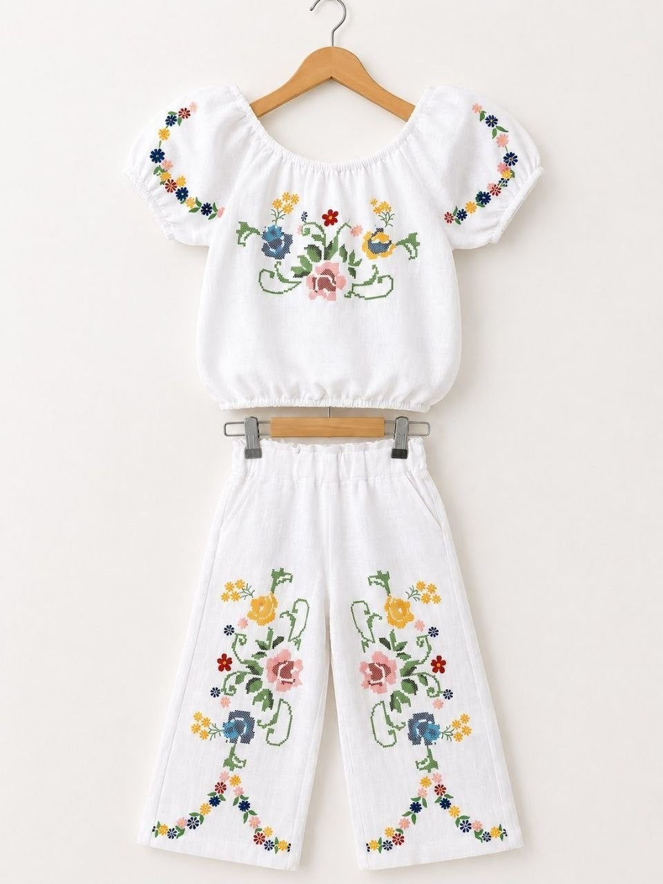 The Nile Luxuries white linen blend off-the-shoulder cropped top with puff sleeves and wide-leg pants, both featuring colorful floral cross-stitch embroidery.