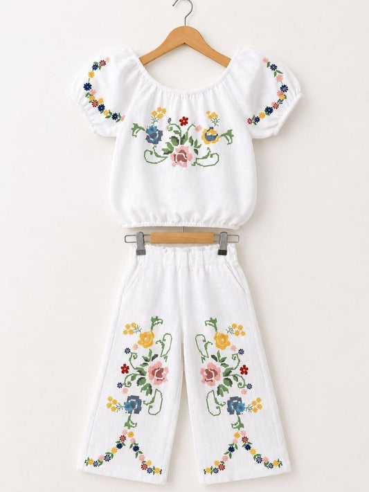 The Nile Luxuries white linen blend off-the-shoulder cropped top with puff sleeves and wide-leg pants, both featuring colorful floral cross-stitch embroidery.