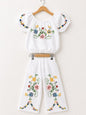 The Nile Luxuries white linen blend off-the-shoulder cropped top with puff sleeves and wide-leg pants, both featuring colorful floral cross-stitch embroidery.