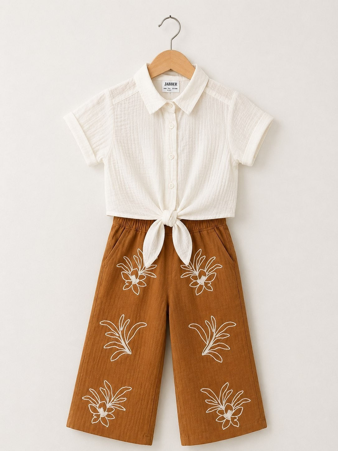 The Nile Luxuries off-white textured short-sleeve collared shirt tied at front, paired with brown wide-leg cropped pants with white floral embroidery.
