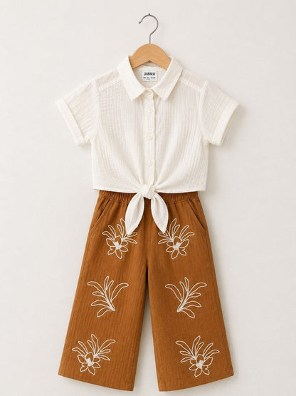 The Nile Luxuries off-white textured short-sleeve collared shirt tied at front, paired with brown wide-leg cropped pants with white floral embroidery.
