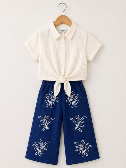 The Nile Luxuries off-white textured short-sleeve collared shirt tied at front, paired with navy blue wide-leg cropped pants with white floral embroidery.