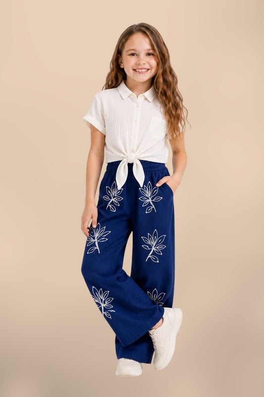 The Nile Luxuries girls' off-white textured shirt tied at front, paired with navy blue wide-leg pants embroidered with white floral pattern.