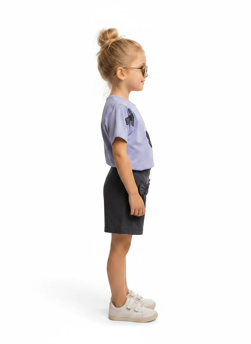The Nile Luxuries girls casual summer outfit with a light purple bow-adorned t-shirt and dark grey shorts, side view.