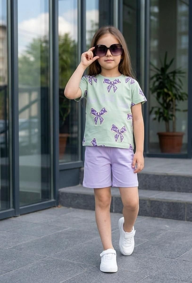 The Nile Luxuries girls' summer set with a sage green bow-print t-shirt and lilac shorts, worn with white sneakers.