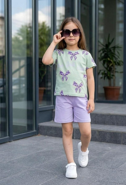 The Nile Luxuries girls' summer set with a sage green bow-print t-shirt and lilac shorts, worn with white sneakers.