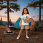 The Nile Luxuries girls' white cotton t-shirt with butterfly graphic and tan shorts. A young girl stands outdoors.