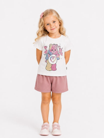Girl wearing The Nile Luxuries white Care Bears graphic cotton t-shirt and mauve shorts against a white background.
