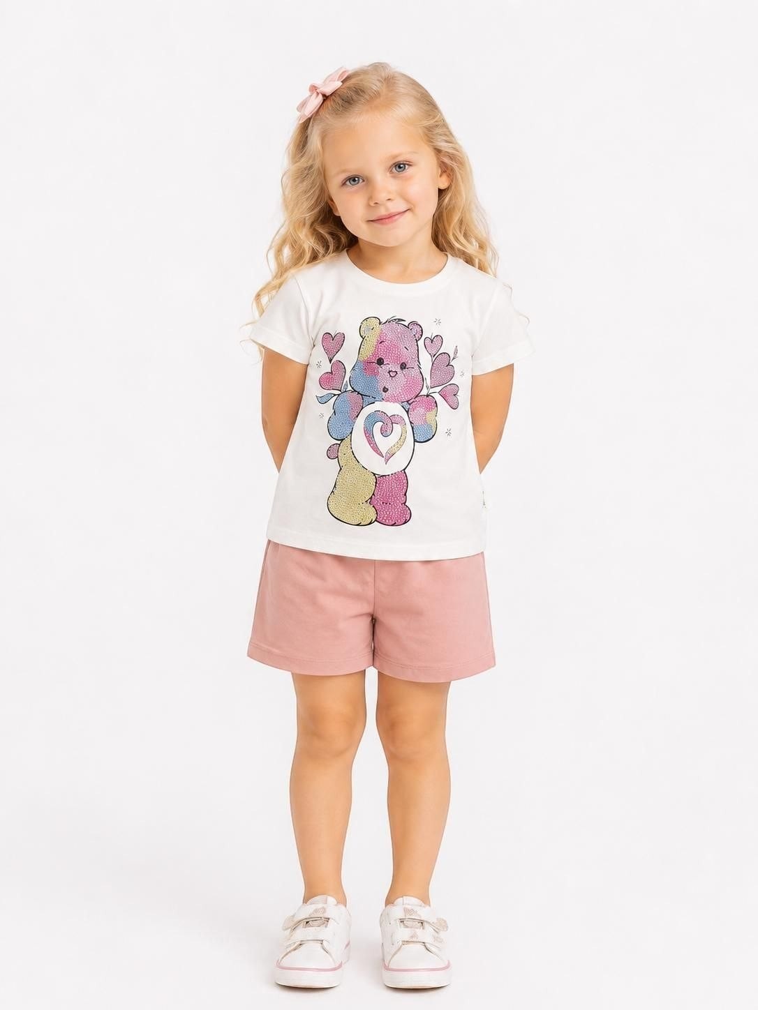 Young girl wearing The Nile Luxuries white Care Bears graphic t-shirt and pink cotton shorts on a white background.