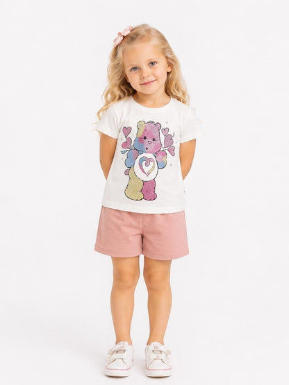 Young girl wearing The Nile Luxuries white Care Bears graphic t-shirt and pink cotton shorts on a white background.