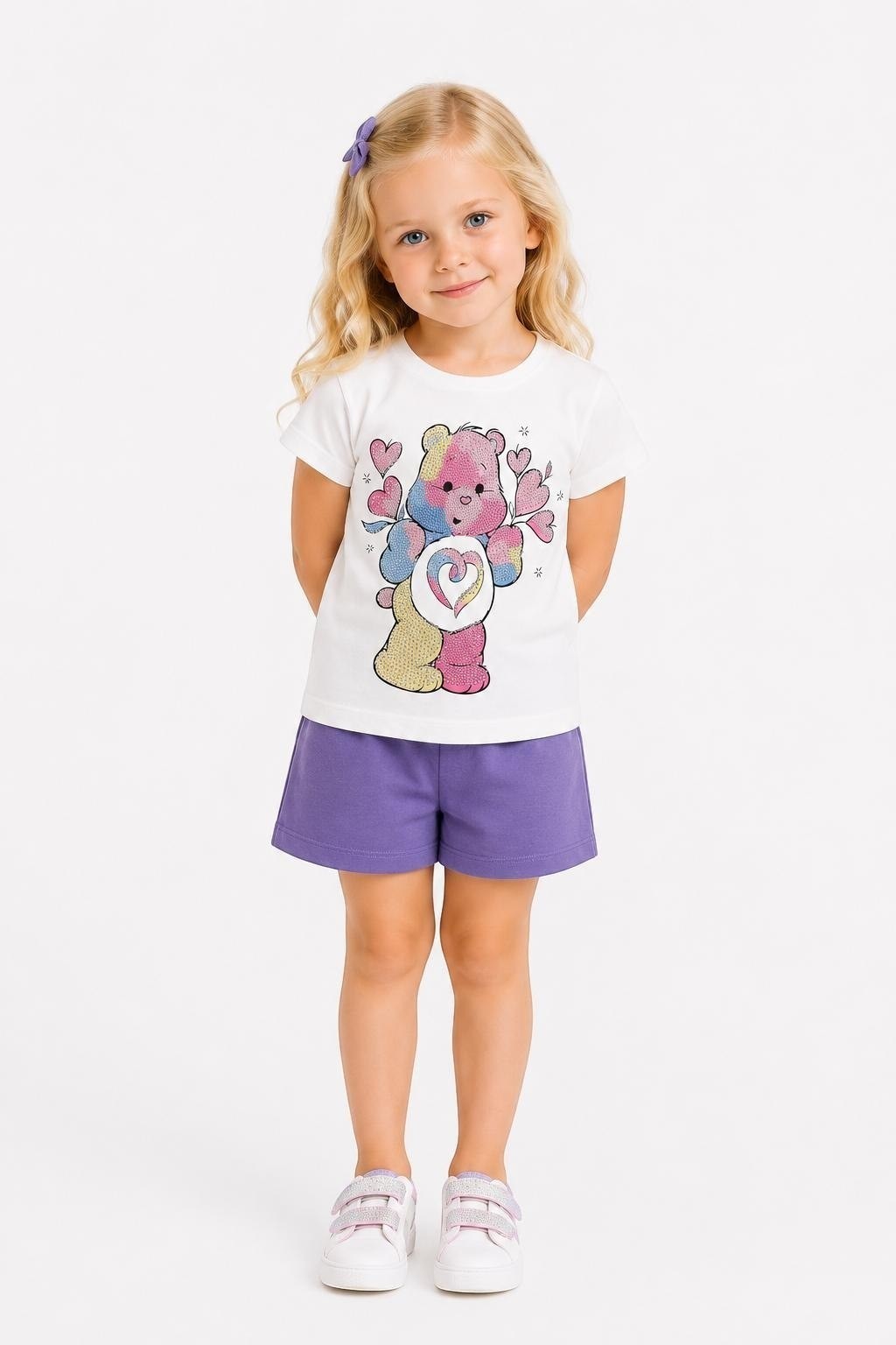 Young girl wearing a The Nile Luxuries white Care Bears graphic tee and purple shorts with white sneakers.