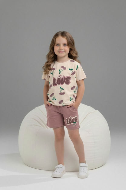 Girl wearing The Nile Luxuries cream cherry-print t-shirt and mauve shorts with LOVE graphic, standing by beanbag.