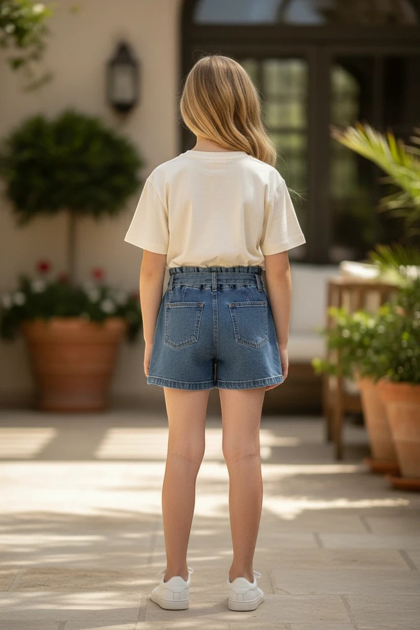 Girl wearing The Nile Luxuries denim shorts with paper-bag waist and cream t-shirt, standing on a patio.