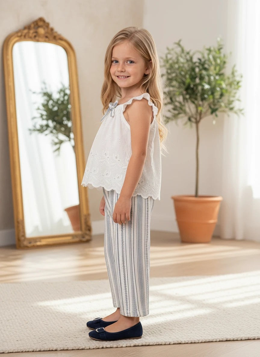 The Nile Luxuries girl wearing a white eyelet lace sleeveless top with flutter sleeves and striped wide-leg trousers.