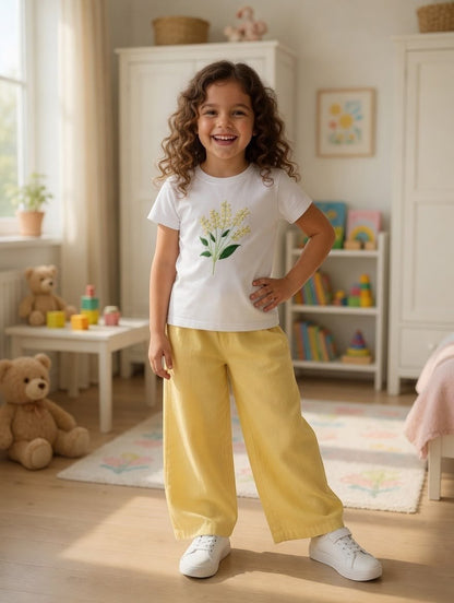 The Nile Luxuries girl wearing a white floral graphic t-shirt and yellow wide-leg pants in a bright bedroom.
