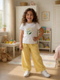 The Nile Luxuries girl wearing a white floral graphic t-shirt and yellow wide-leg pants in a bright bedroom.