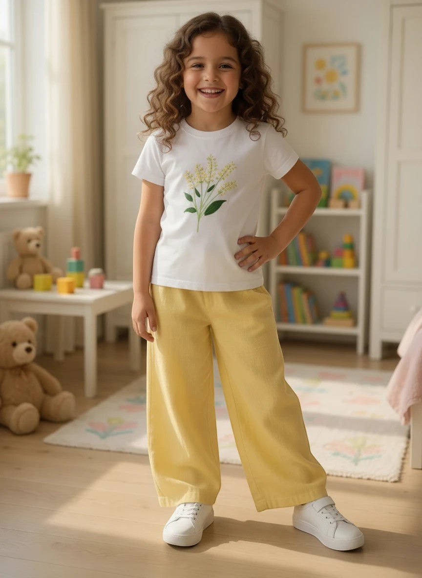 A young girl wearing The Nile Luxuries white floral graphic t-shirt and yellow wide-leg pants in a bright bedroom.