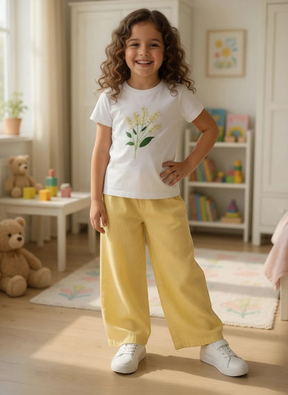 A young girl wearing The Nile Luxuries white floral graphic t-shirt and yellow wide-leg pants in a bright bedroom.