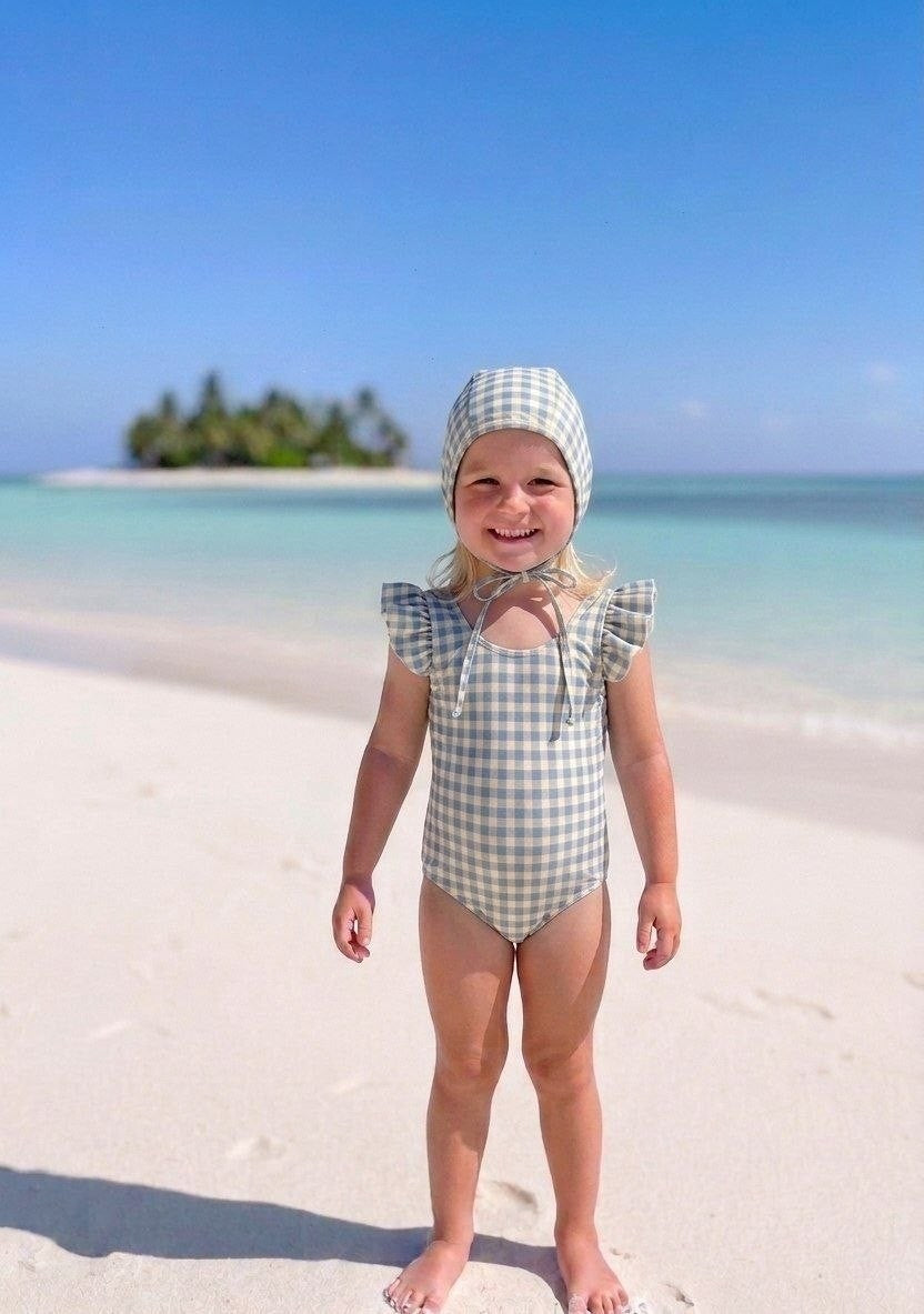 The Nile Luxuries girls' blue and cream gingham ruffle sleeve swimsuit and matching bonnet on a beach.