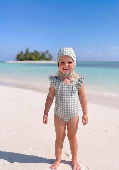 The Nile Luxuries girls' blue and cream gingham ruffle sleeve swimsuit and matching bonnet on a beach.