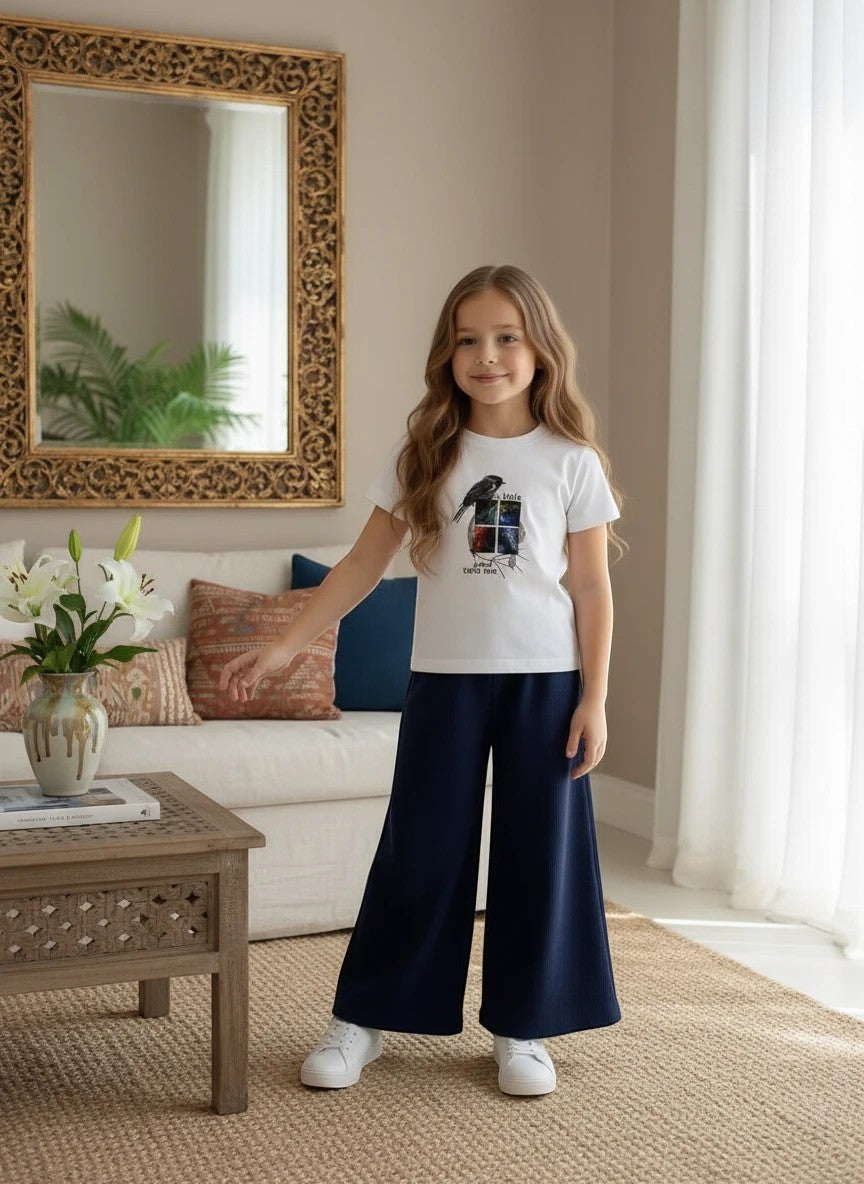 Girl wearing The Nile Luxuries white bird graphic t-shirt and navy blue ribbed wide-leg trousers in a living room.