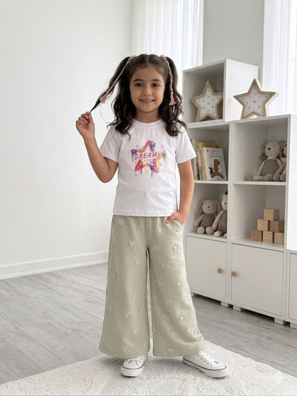 A young girl wearing a The Nile Luxuries white Dreams graphic t-shirt and sage green wide-leg star-patterned pants.