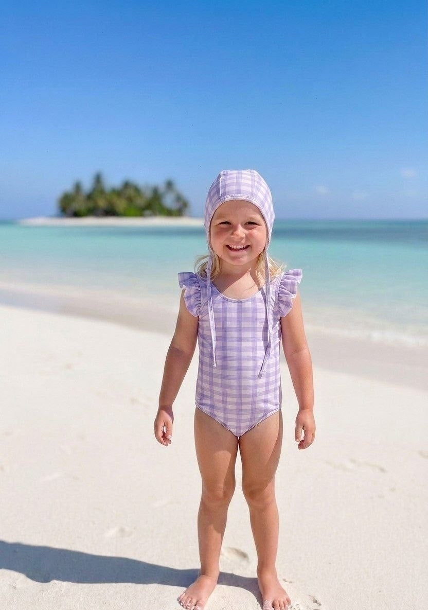 The Nile Luxuries girls' lavender gingham swimsuit with ruffle sleeves and matching bonnet on a beach.