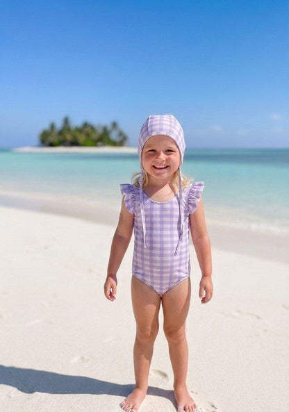 The Nile Luxuries girls' lavender gingham swimsuit with ruffle sleeves and matching bonnet on a beach.