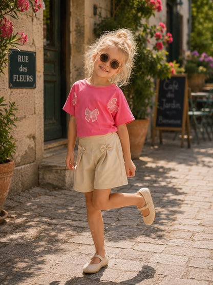 The Nile Luxuries girls summer outfit with a pink bow-adorned t-shirt and beige shorts worn by a smiling young girl.