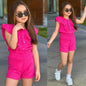A young girl wearing a bright pink The Nile Luxuries ruffle top and shorts set with white sneakers and sunglasses.