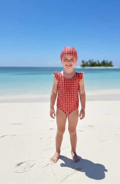 The Nile Luxuries girls red gingham ruffle sleeve swimsuit and matching bonnet on a beach.